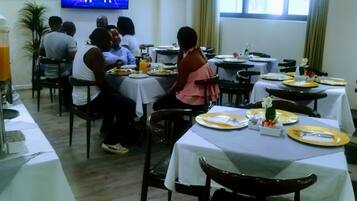 A restaurant with multiple tables, some occupied by diners. A television is mounted on the wall displaying a sports event.