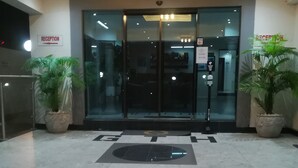 Ein Hotel-Empfangsbereich mit einem 'WELCOME'-Schild auf dem Boden, Glastüren, Topfpflanzen und einer Deckenleuchte.