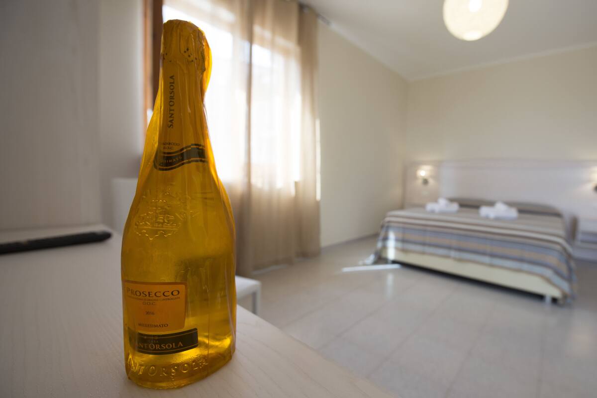 Prosecco-pullo hotellihuoneen pöydällä, huoneessa on myös sänky ja yöpöytä.