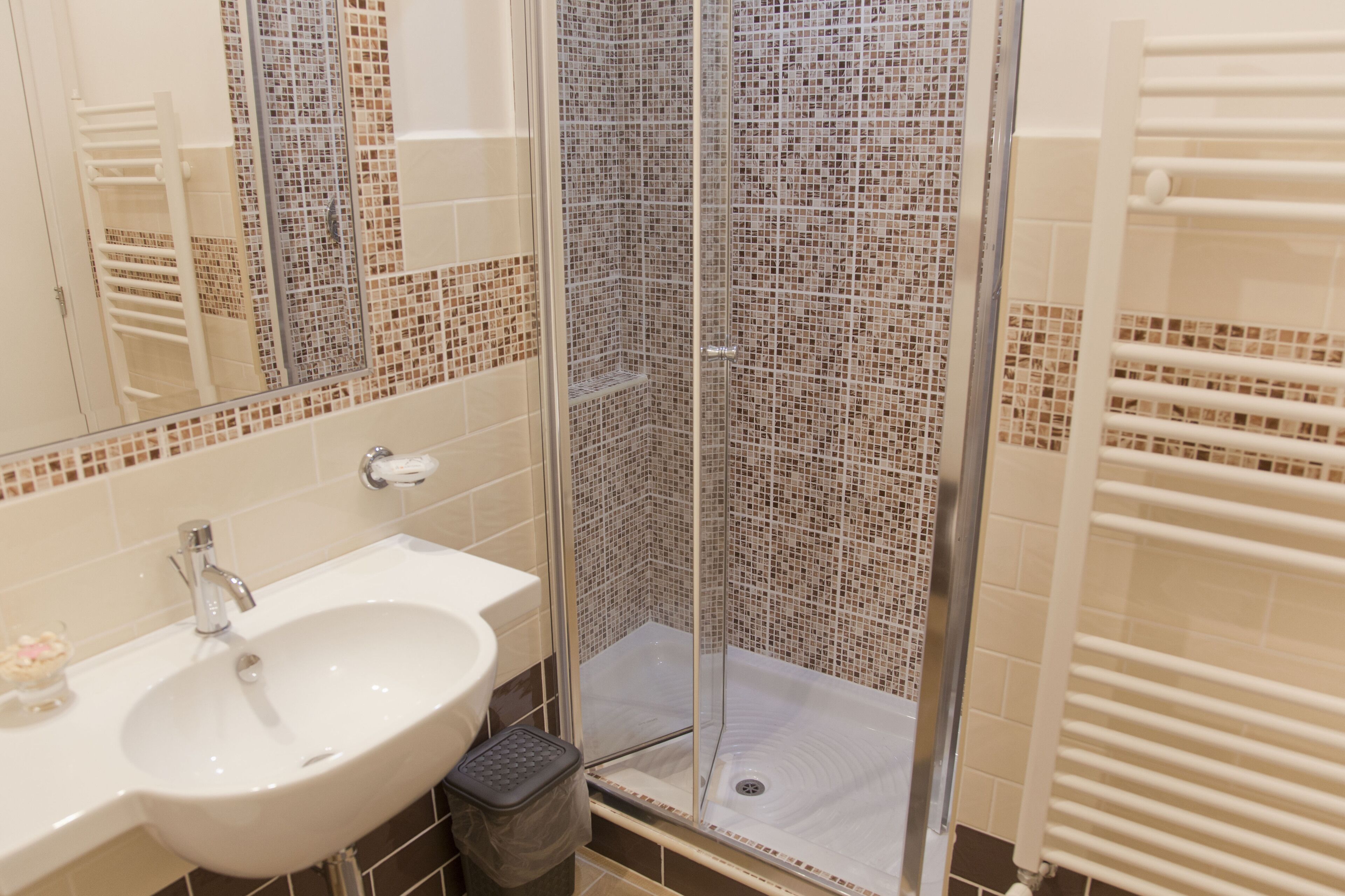 Un baño con lavamanos blanco, espejo y ducha con mampara de vidrio y mosaicos.