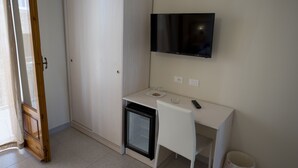 Een compacte kamer met een wit bureau, een kleine koelkast, een aan de muur bevestigde televisie en een garderobe.
