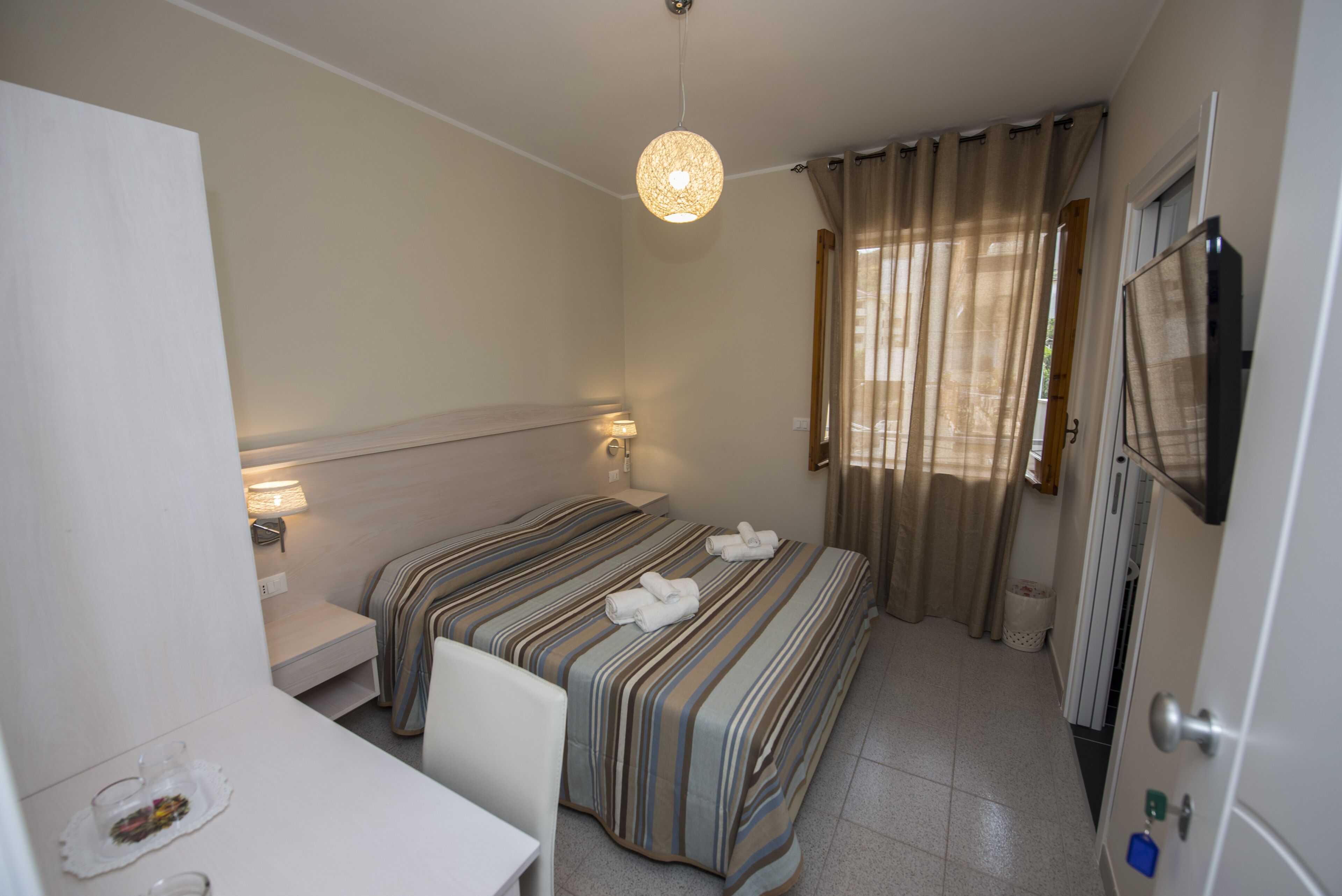 Una habitación de hotel con cama, mesitas de noche, un escritorio, televisión y cortinas.