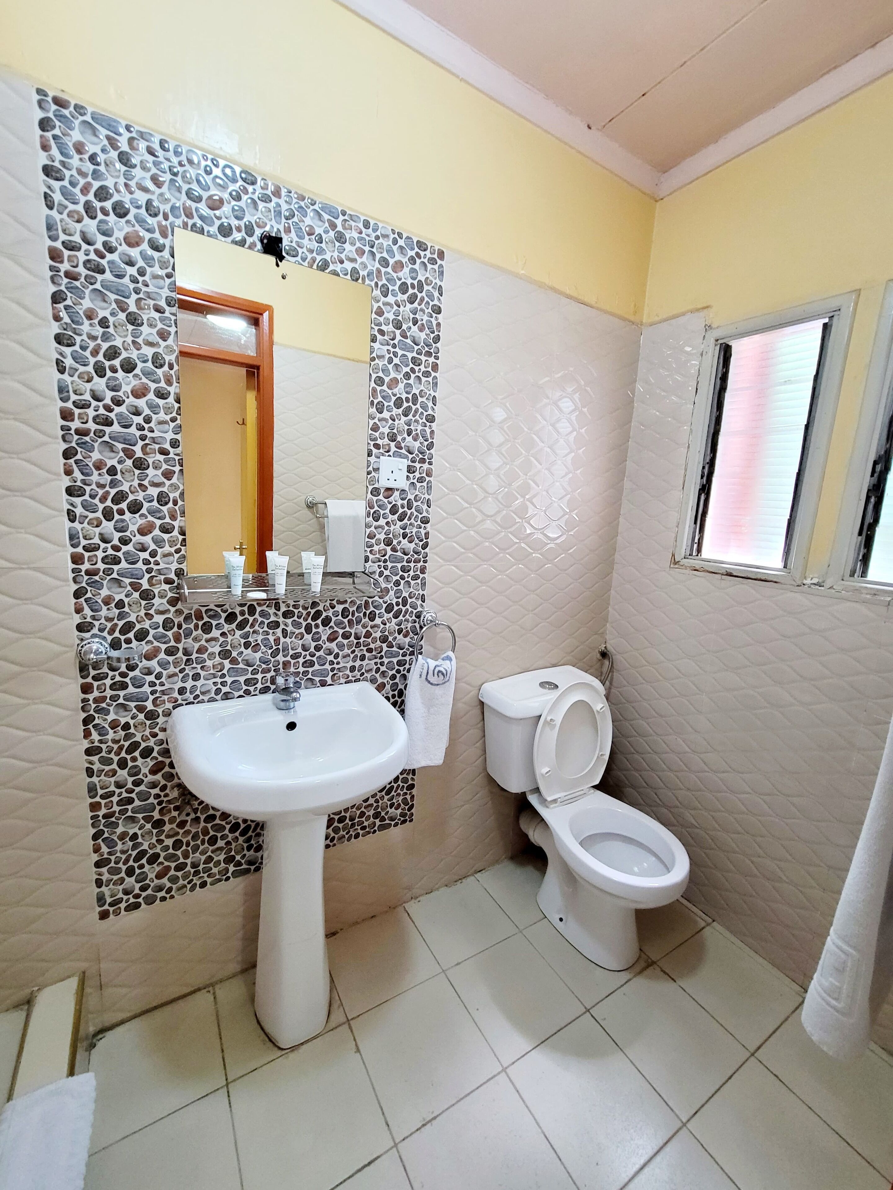 A bathroom with a stone mosaic backsplash, a white sink, a toilet, and a window with blinds.