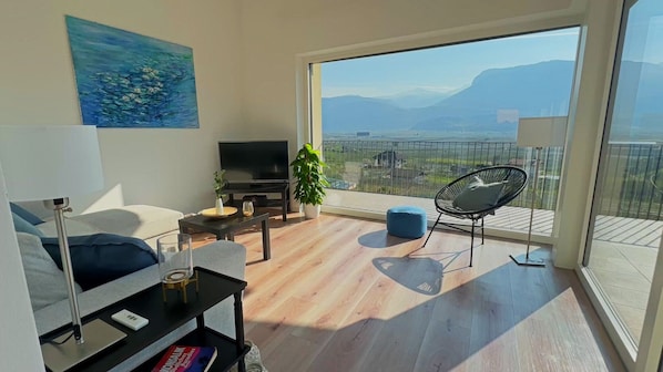 A modern living room with a TV, seating, a plant, and a balcony with a city view.