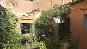 A balcony with a variety of plants and flowers, a window, and a building in the background. - Chambres d'Hôtes de Fleurus (Pezenas)