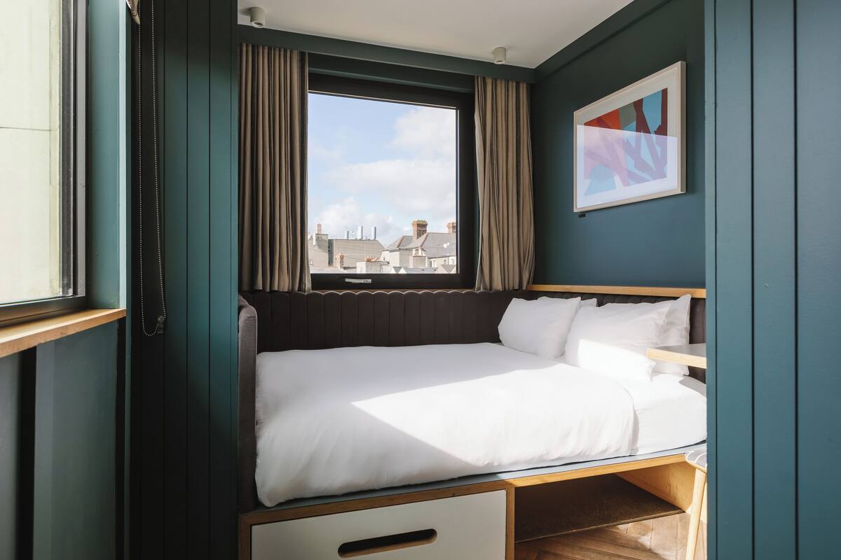 A compact, well-lit room with a bed, a window with curtains, and a framed abstract painting on the wall.