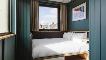 A compact, well-lit room with a bed, a window with curtains, and a framed abstract painting on the wall.