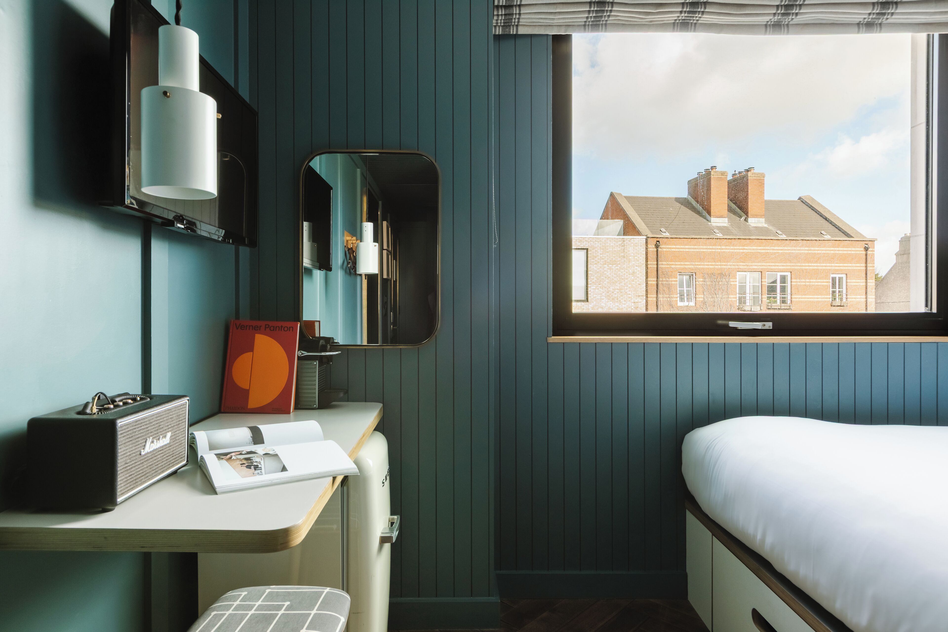 A room with a bed, a desk with a mirror, a radiator, and a window with a view of a building.