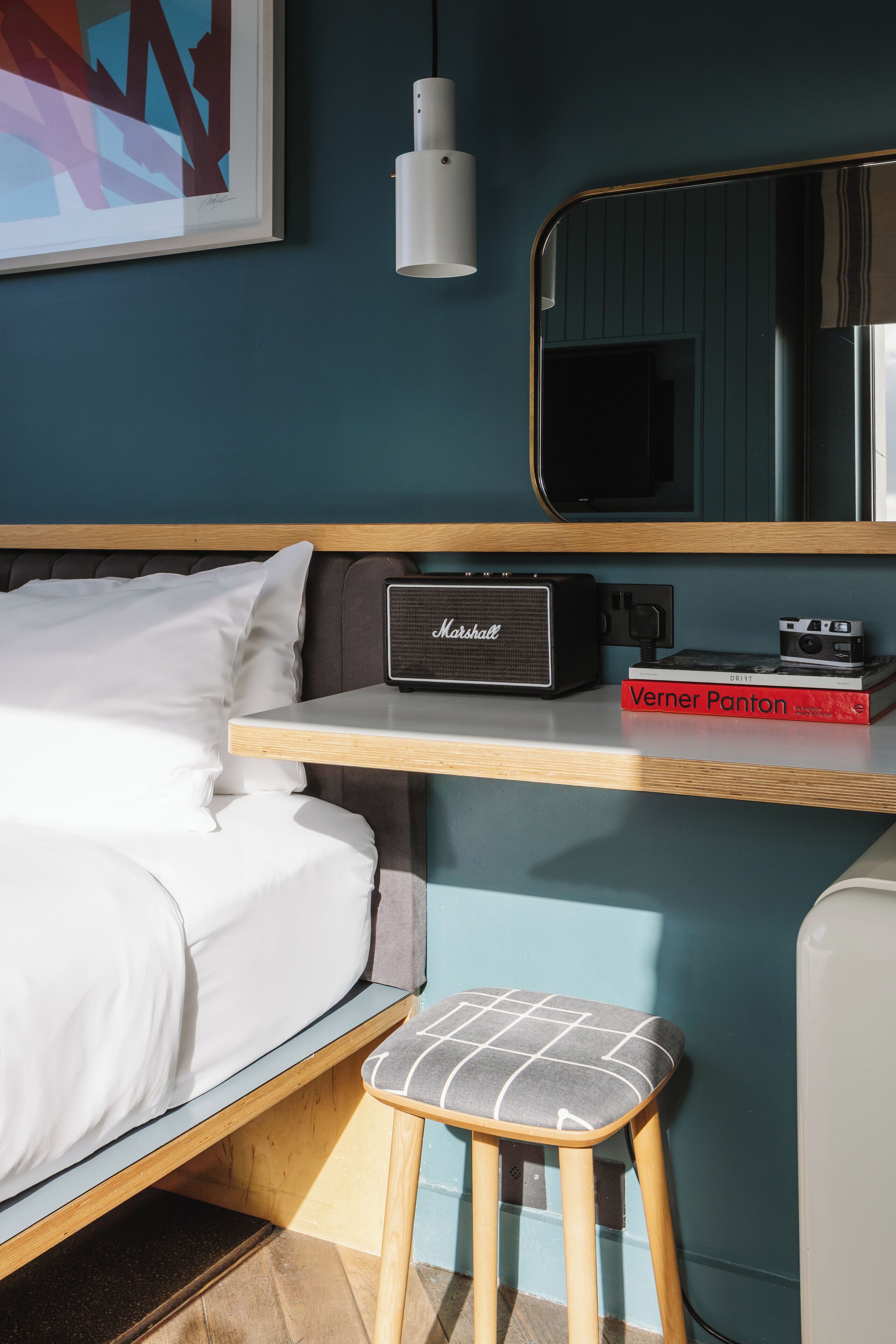 A hotel room with a bed, a small stool, a Marshall amplifier, a book titled 'Verner Panton', a mirror, and a wall-mounted light fixture.