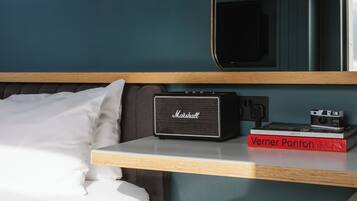 A hotel room with a bed, a small stool, a Marshall amplifier, a book titled 'Verner Panton', a mirror, and a wall-mounted light fixture.