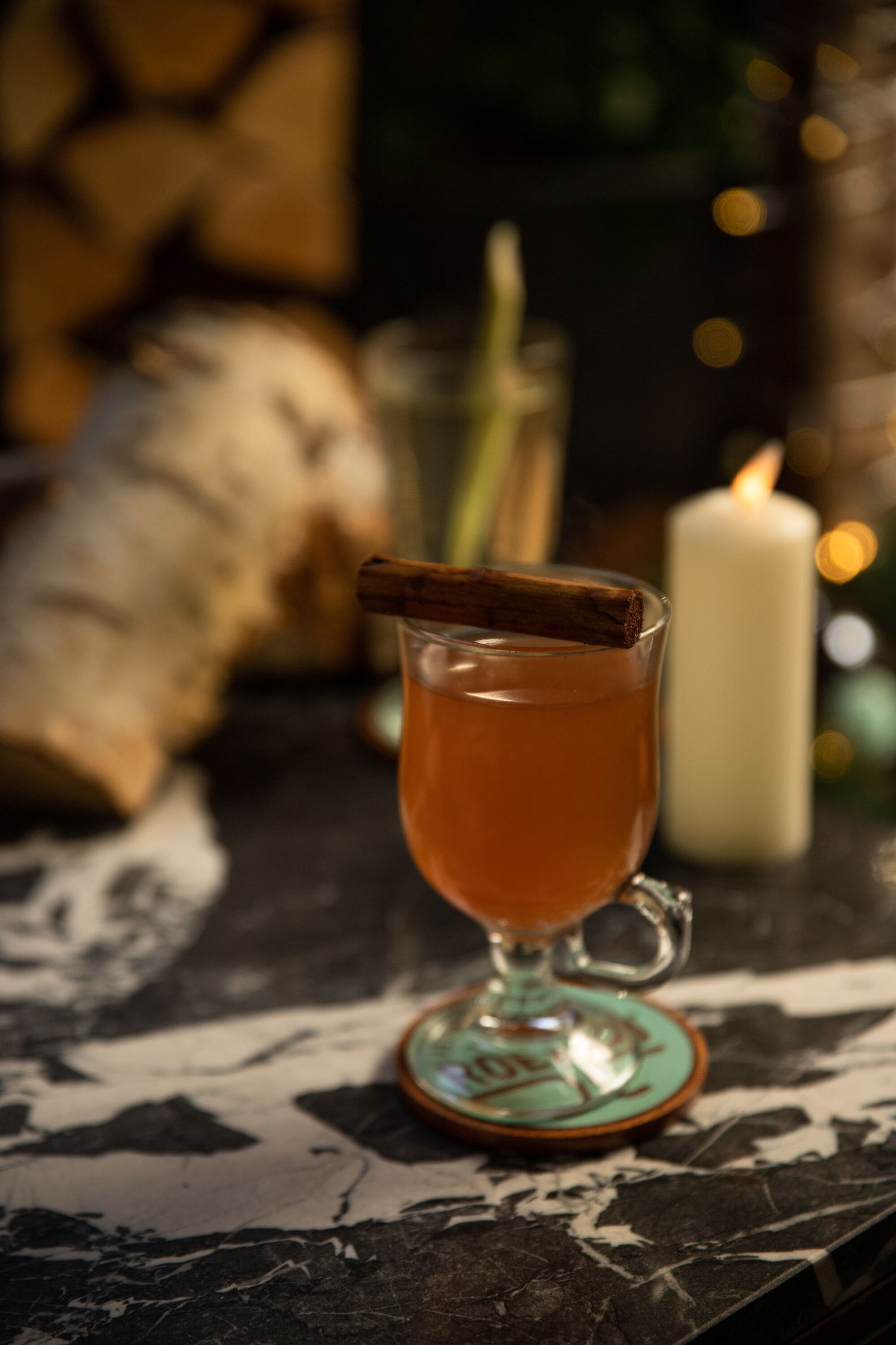 A glass of amber liquid with a cinnamon stick, a candle, and a decorative plate on a marble surface.