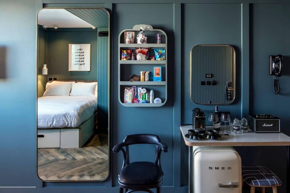 A hotel room with a bed, a desk with a coffee maker, a chair, a minibar, and a mirror.