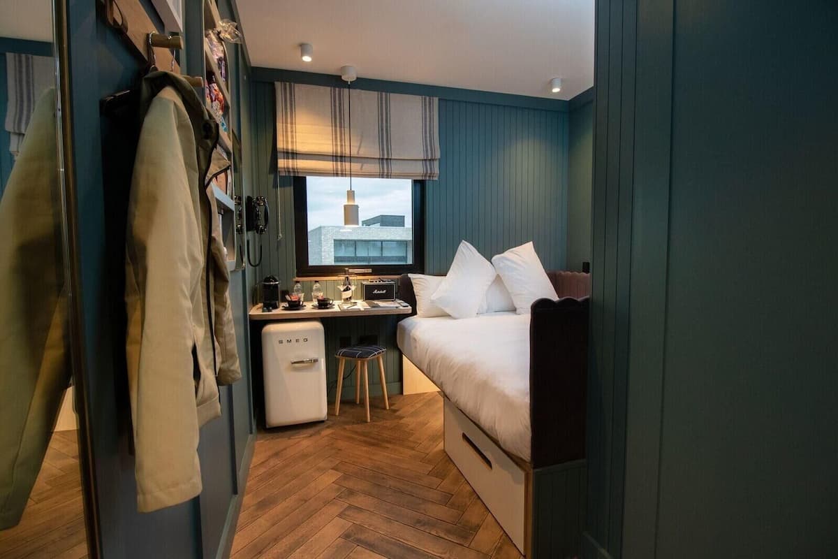 A compact hotel room with a bed, a desk, a chair, and a small kitchenette.