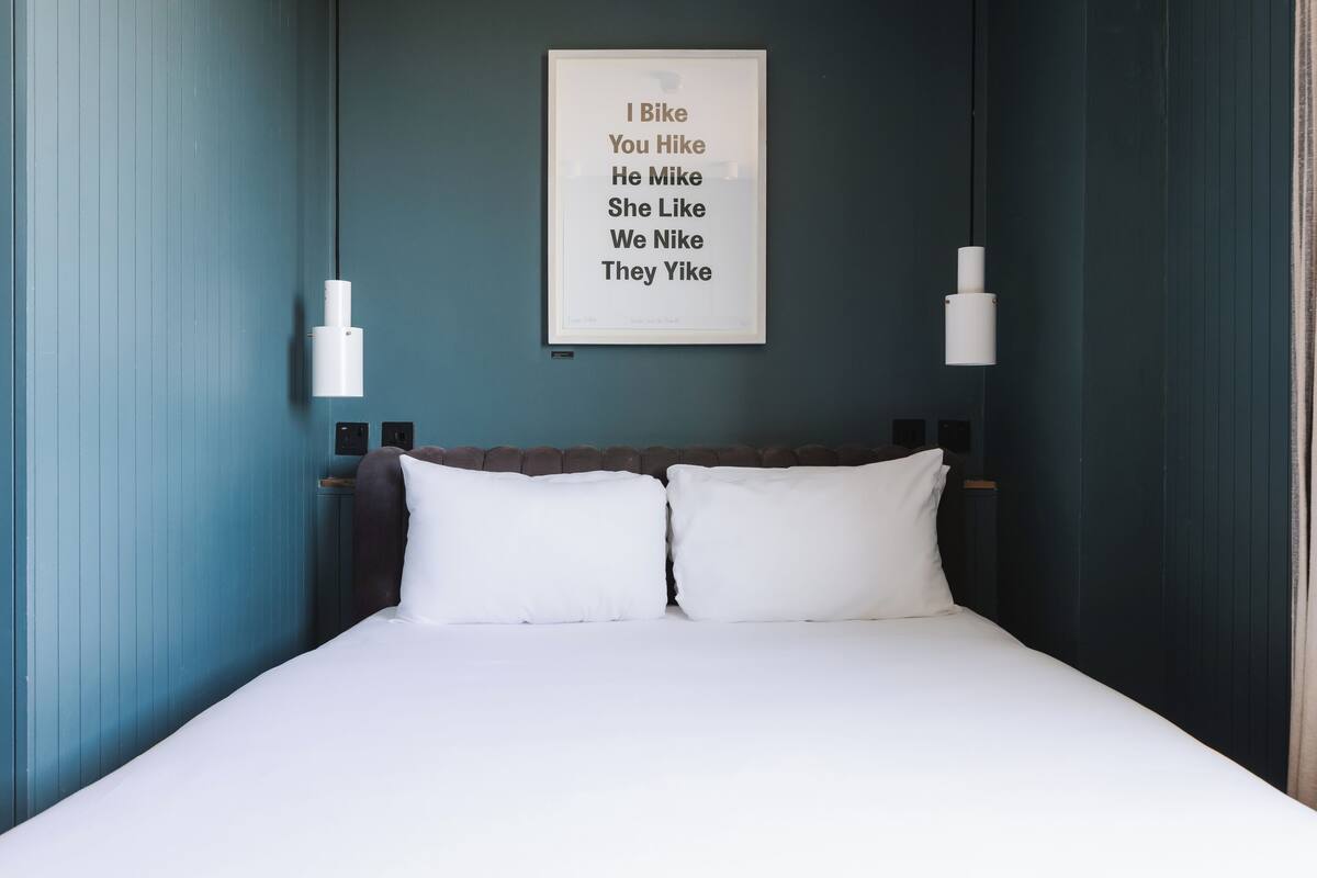 A neatly made bed with white bedding, a framed text on the wall, and two pendant lights.
