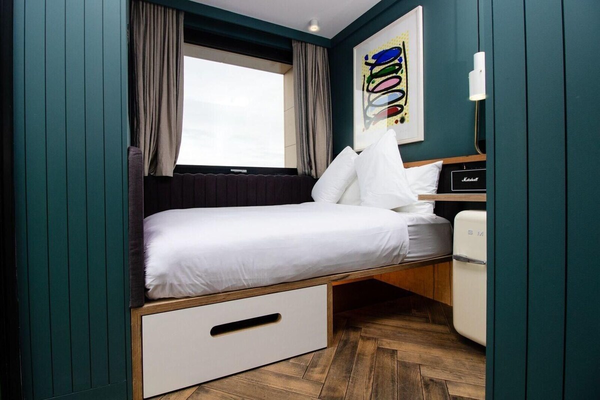 A small, neatly arranged room with a bed, a desk, a chair, and a window with curtains.