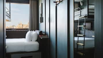 A compact hotel room with a bunk bed, a sofa bed, a bedside table with a lamp, and a view of the cityscape through a large window.