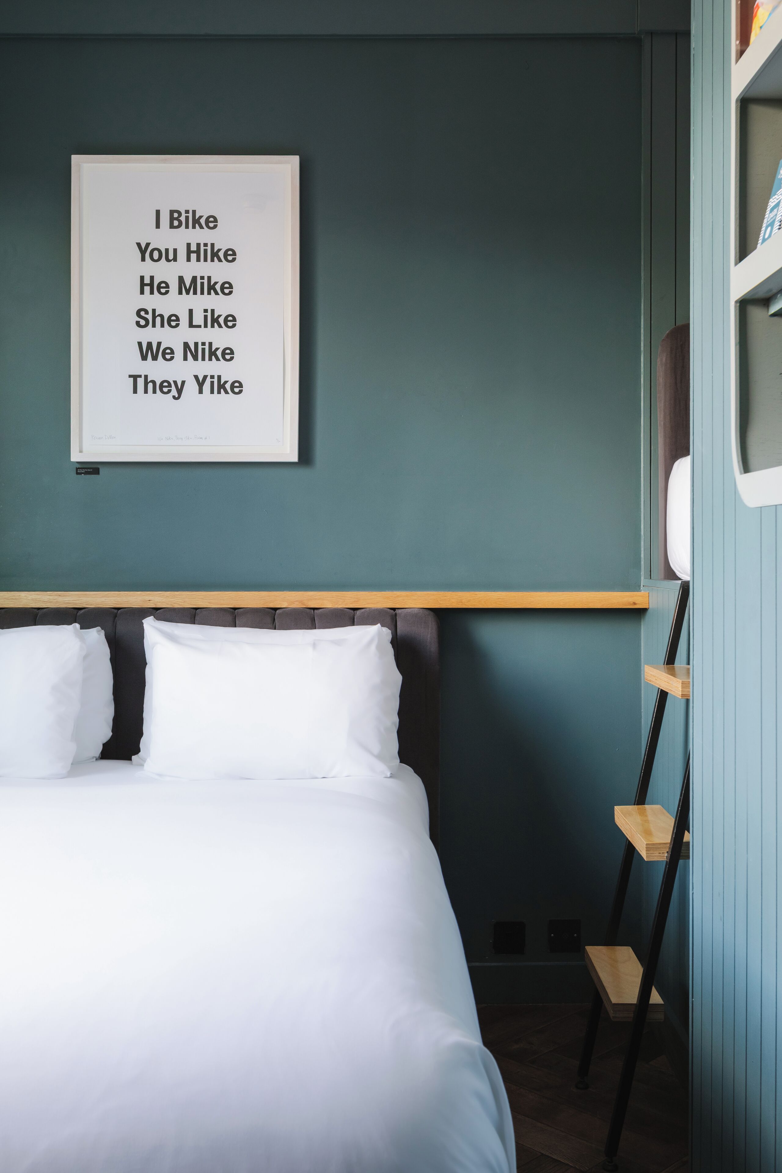 A neatly made bed with white linens, a framed quote on the wall, and a wooden shelf unit beside the bed.