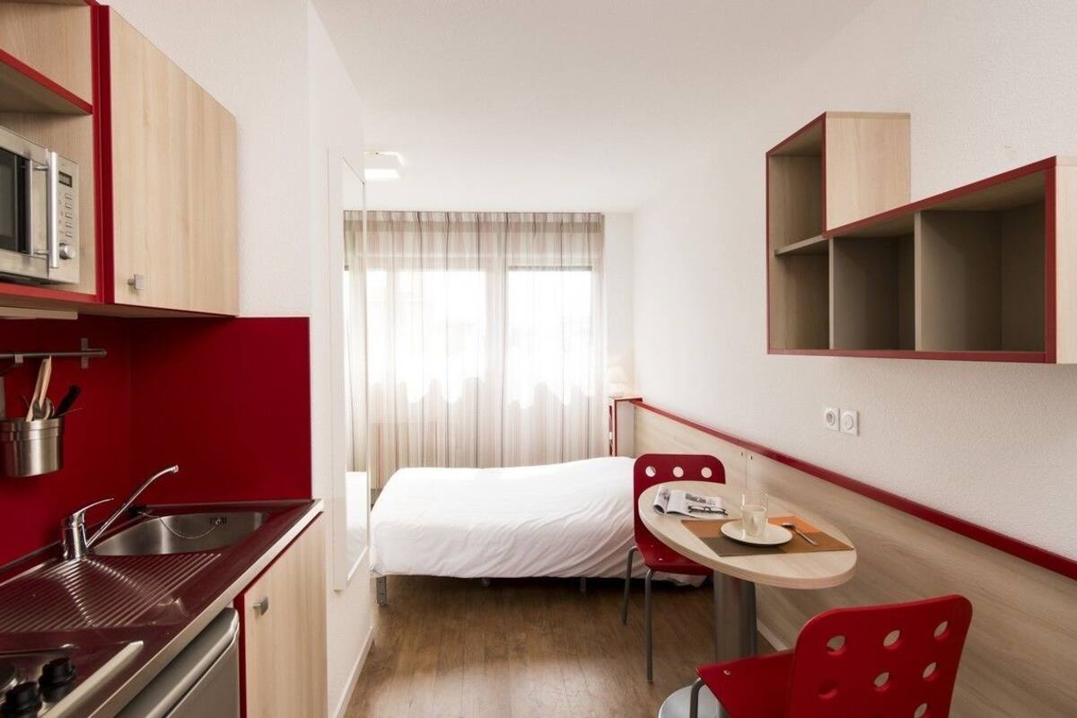 A compact hotel room with a kitchenette, a bed, a dining area, and a window with sheer curtains.