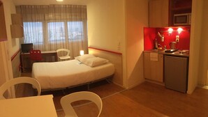 Ett kompakt hotellrum med en enkel säng, ett skrivbord, en stol, ett litet pentry och ett fönster med gardiner.
