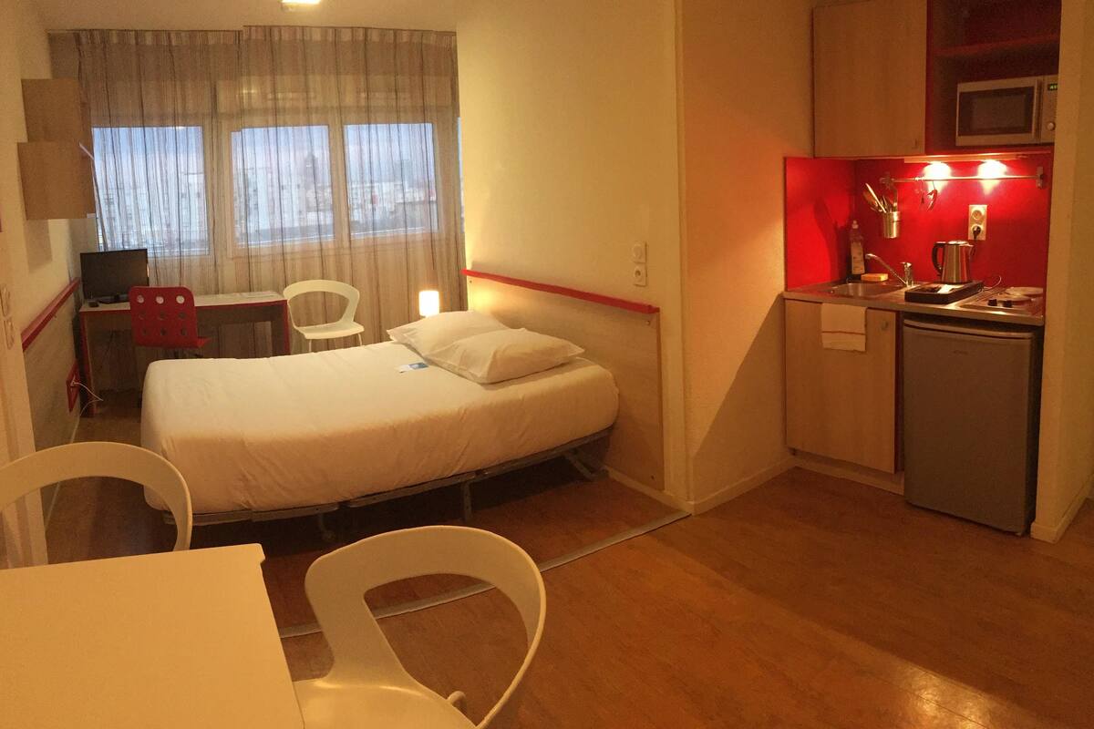 A compact hotel room with a single bed, a desk, a chair, a small kitchenette, and a window with curtains.