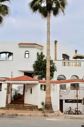 A white building with a red-tiled roof, a palm tree in front, and a bicycle parked by the gate.