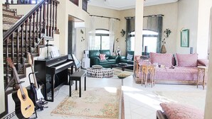 Un salón amplio con un piano, un sofá verde y un sofá rosa. Se ve una escalera con barandillas de madera al fondo.