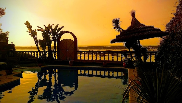 Una piscina con vista al atardecer, una estructura de techo de paja y una barandilla sobre el agua.