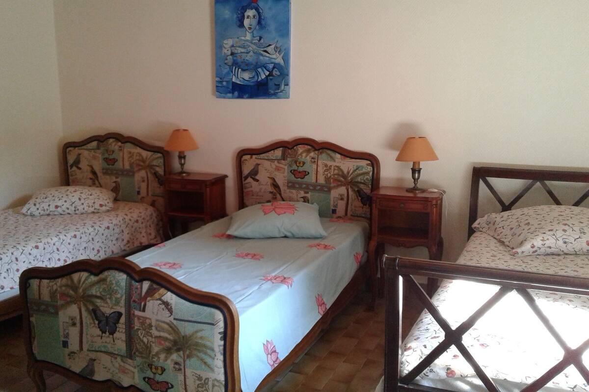 A room with three single beds, each with a wooden headboard and floral-patterned bedding. There are two bedside tables with lamps, and a painting hangs on the wall above the beds.