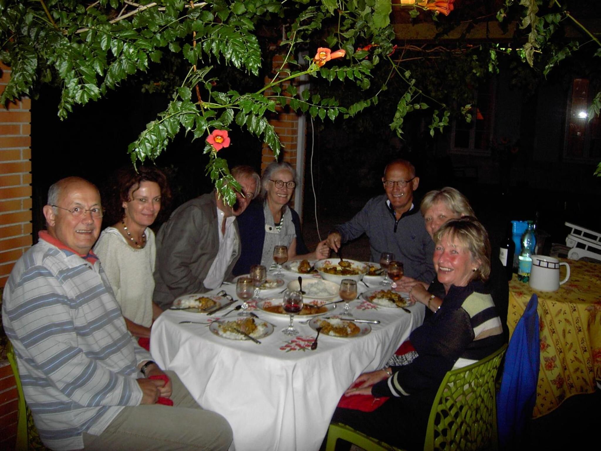 A group of people dining at an outdoor table with a white tablecloth, surrounded by greenery and a brick wall.