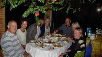 A group of people dining at an outdoor table with a white tablecloth, surrounded by greenery and a brick wall.