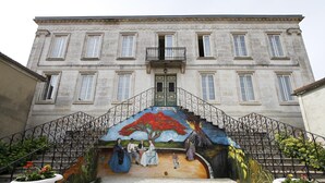 Un edificio con una escalera que lleva a una puerta, rodeado por un mural con figuras y un árbol.