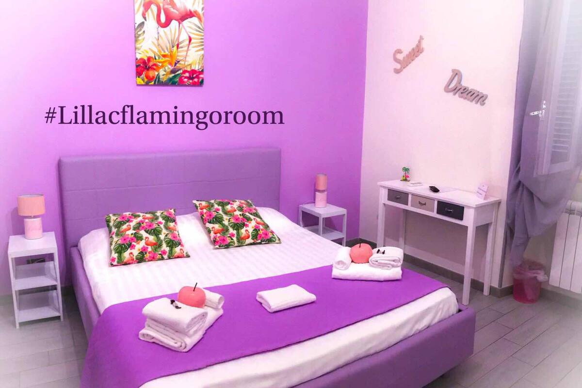 A room with a purple bed, white pillows, and a pink flamingo decoration. There is a small table with a lamp and a trash can.