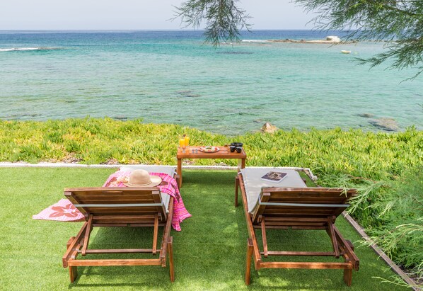 Two wooden chairs and a table set up on a grassy area overlooking a clear blue sea with a rocky shoreline in the background. - Niki Savvas Studio & Suites (Milos)
