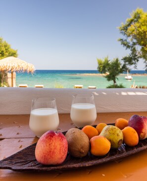 A table with two glasses of milk and a variety of fruits including peaches and kiwis, set against a backdrop of a beach and ocean view. - Niki Savvas Studio & Suites (Milos)