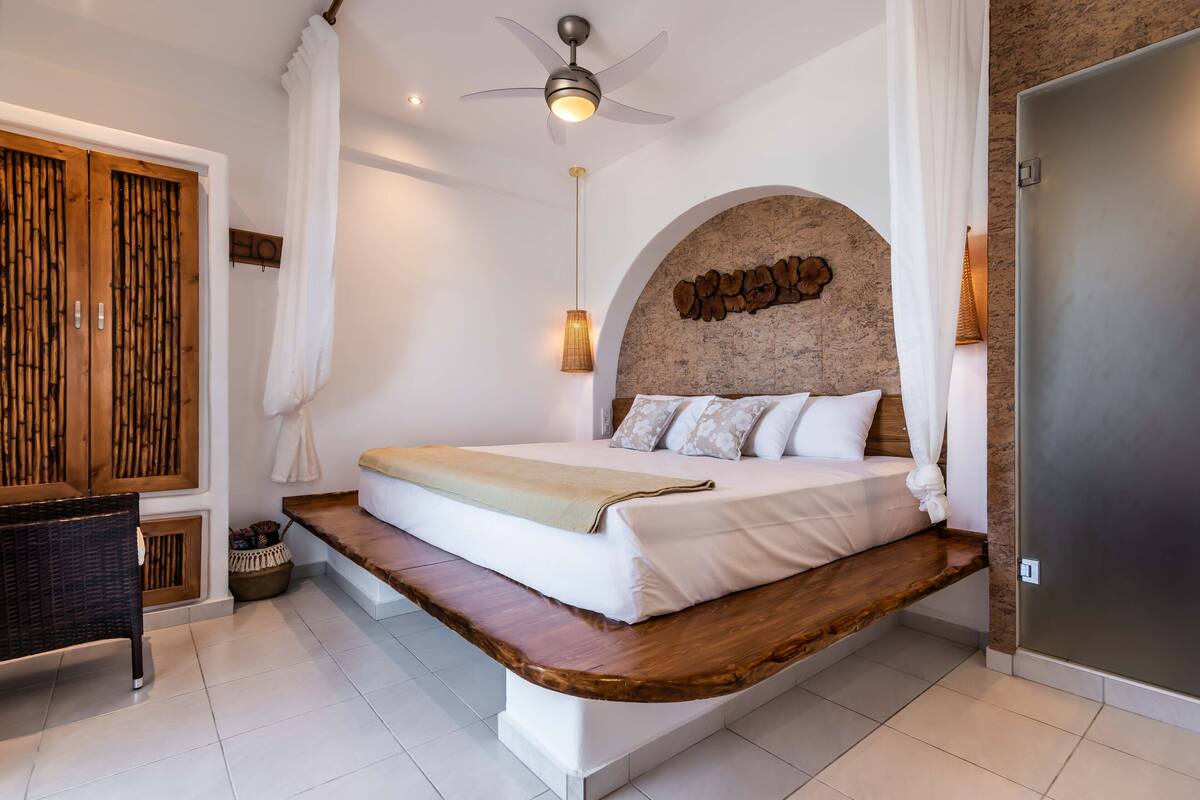 A modern bedroom with a bed, wooden headboard, ceiling fan, and a large mirror.