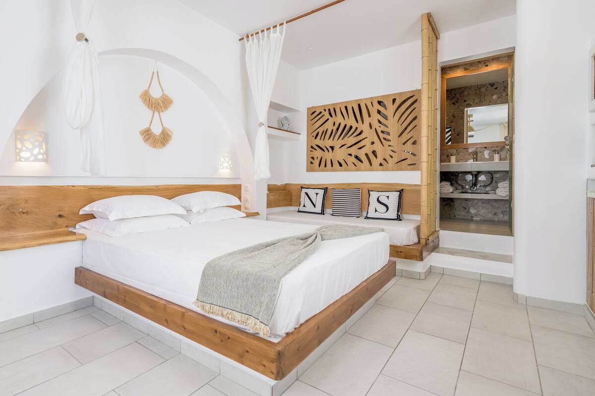 A bedroom with a wooden bed frame, white bedding, a canopy bed, a decorative wall panel, and a bathroom visible through an open door.