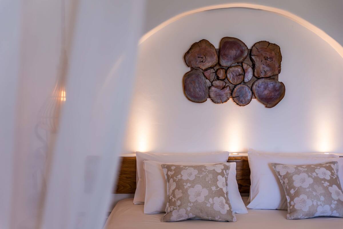 A bedroom with a bed, floral-patterned pillows, a wooden headboard, and a wall-mounted wooden art piece.