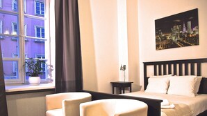 A hotel room with a bed, two armchairs, a nightstand, a window with curtains, and a cityscape painting on the wall. - Old Town Snug (Warsaw)