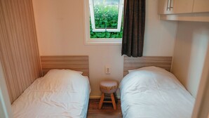 Quarto pequeno com duas camas de solteiro, um banquinho, janela com vista para vegetação e um armário branco.