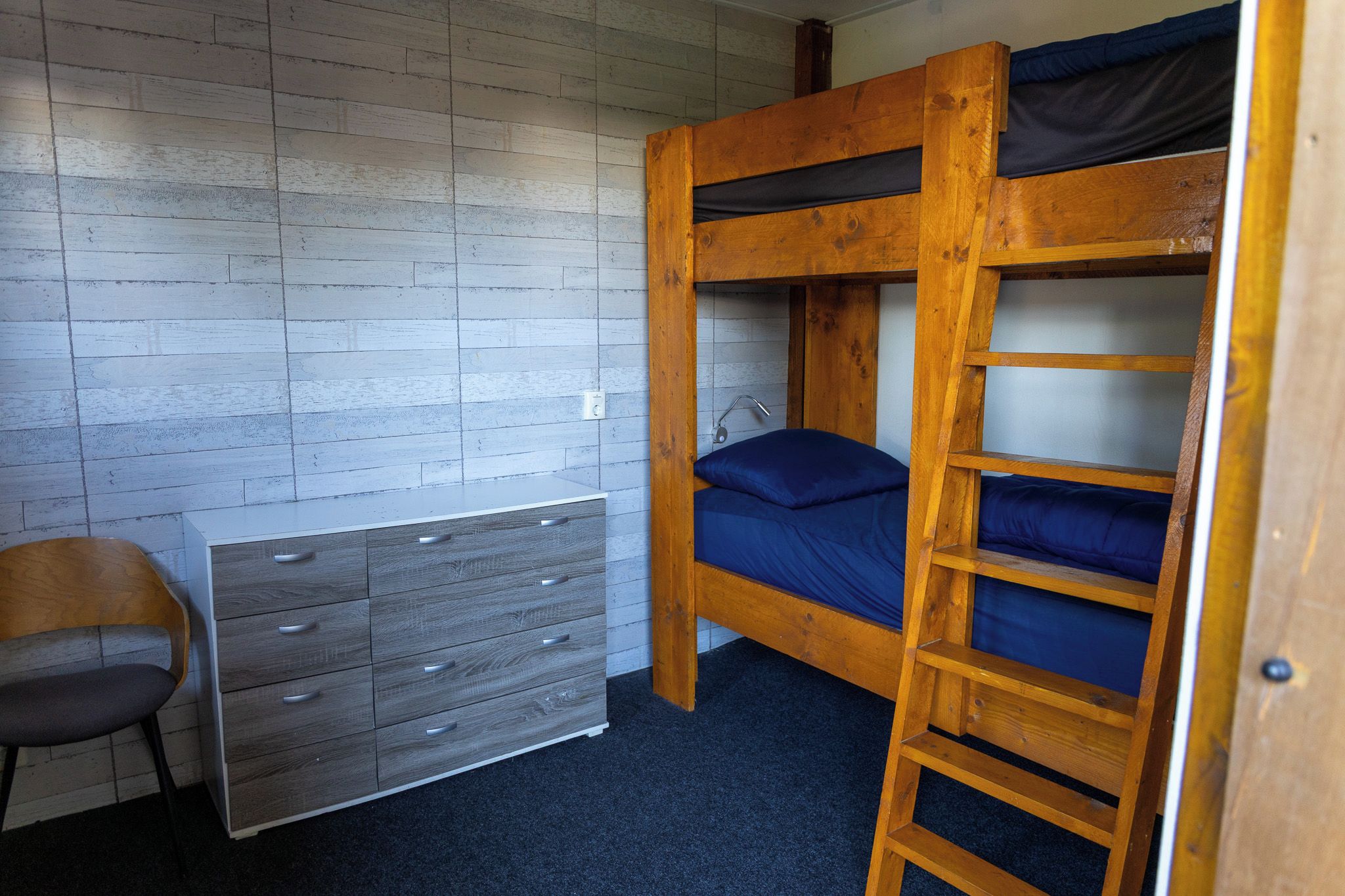 Un llit amb lliteres amb una escala, un escriptori amb calaixos i una cadira en una habitació amb paret de fusta.