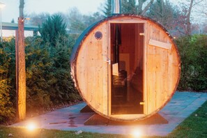 Une structure cylindrique en bois, dotée d’une porte, qui pourrait être un sauna ou un jacuzzi, installée sur un chemin pavé, le tout entouré de verdure.