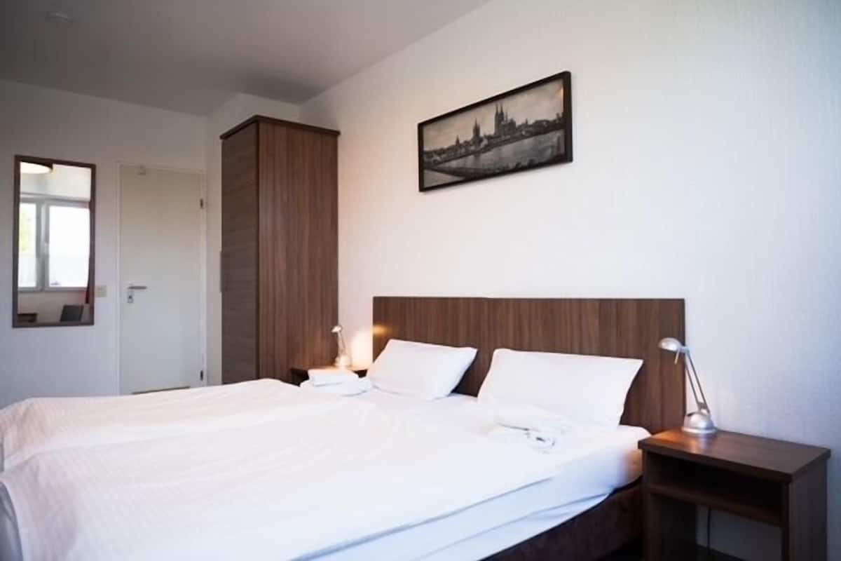 Quarto de hotel com uma cama grande, cabeceira de madeira, duas mesas de cabeceira com abajures, um quadro com vista da cidade na parede e uma porta à esquerda.