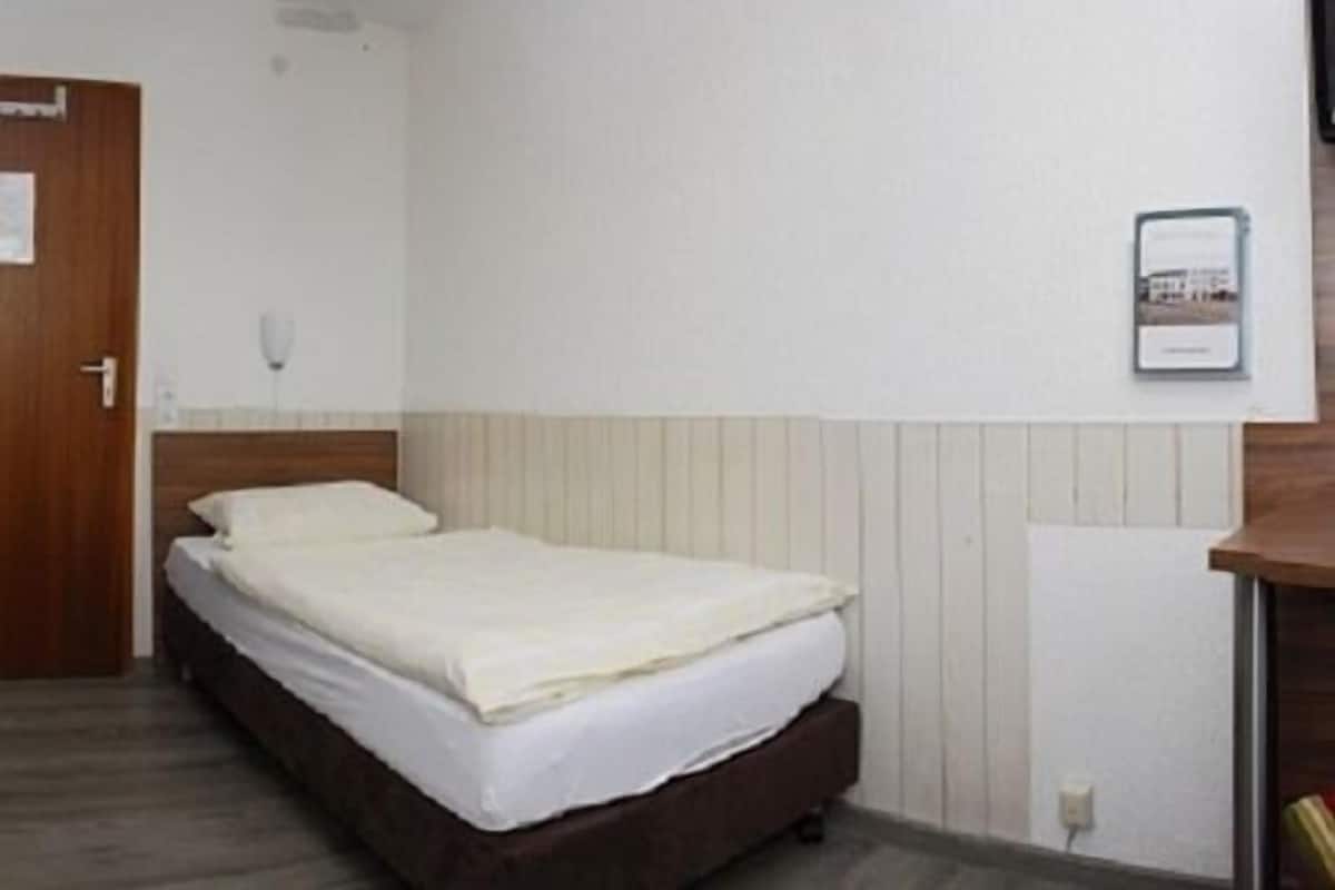 Quarto individual com uma cama de solteiro, cabeceira de madeira, mesa de cabeceira e telefone fixado na parede.