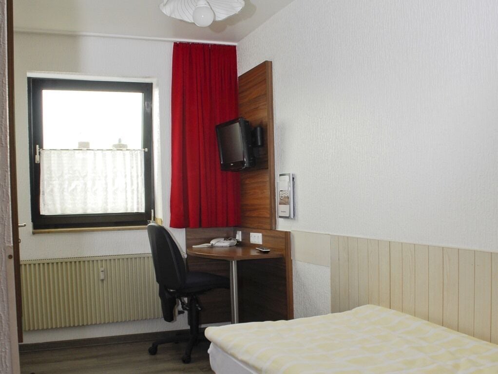 A hotel room with a bed, a desk with a chair, a television mounted on the wall, and a window with red curtains.