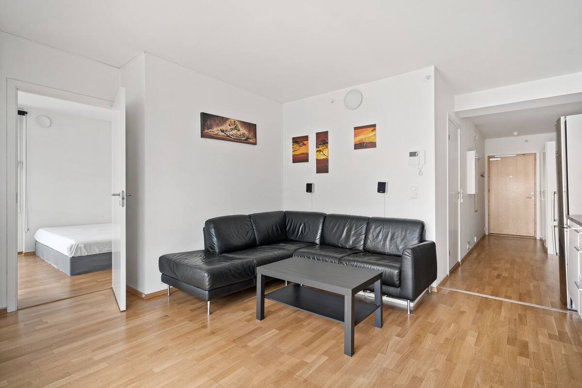 A modern living room with a black leather sectional sofa, a grey coffee table, and a wooden floor. There are three framed artworks on the wall and a door leading to another room.