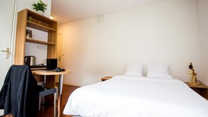 A hotel room with a bed, a desk with a laptop, a chair, a coat rack, and a shelf with a plant and a jar. - Studilodge - Zola Park (Villeurbanne)