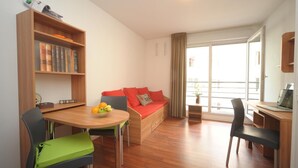 A modern living room with a wooden dining table, a sofa with red cushions, a bookshelf, and a laptop on a desk. - Studilodge - Zola Park (Villeurbanne)