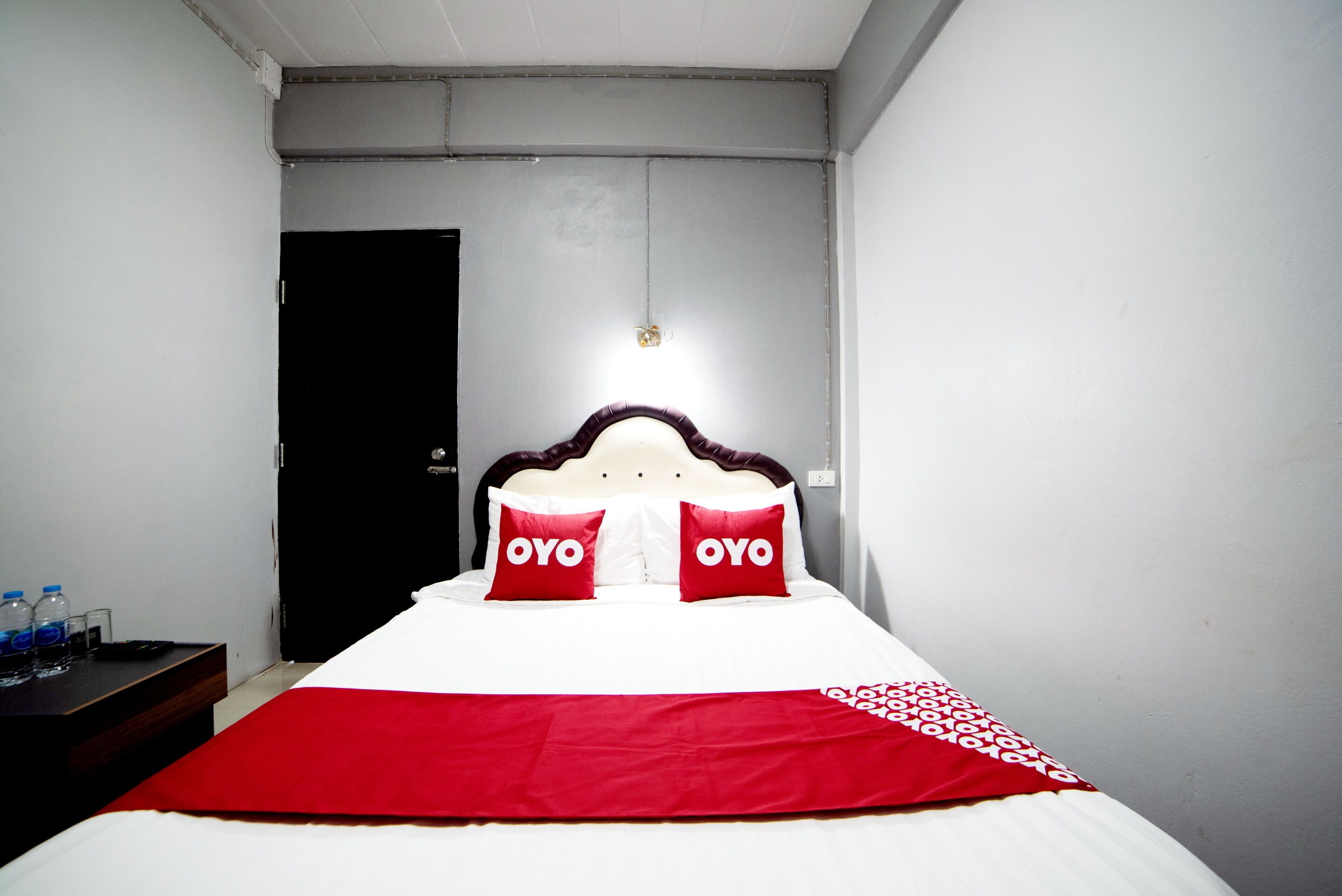 Una cama con cabecera, dos almohadas con la marca 'OYO', una colcha roja y una mesita de noche con botellas de agua.