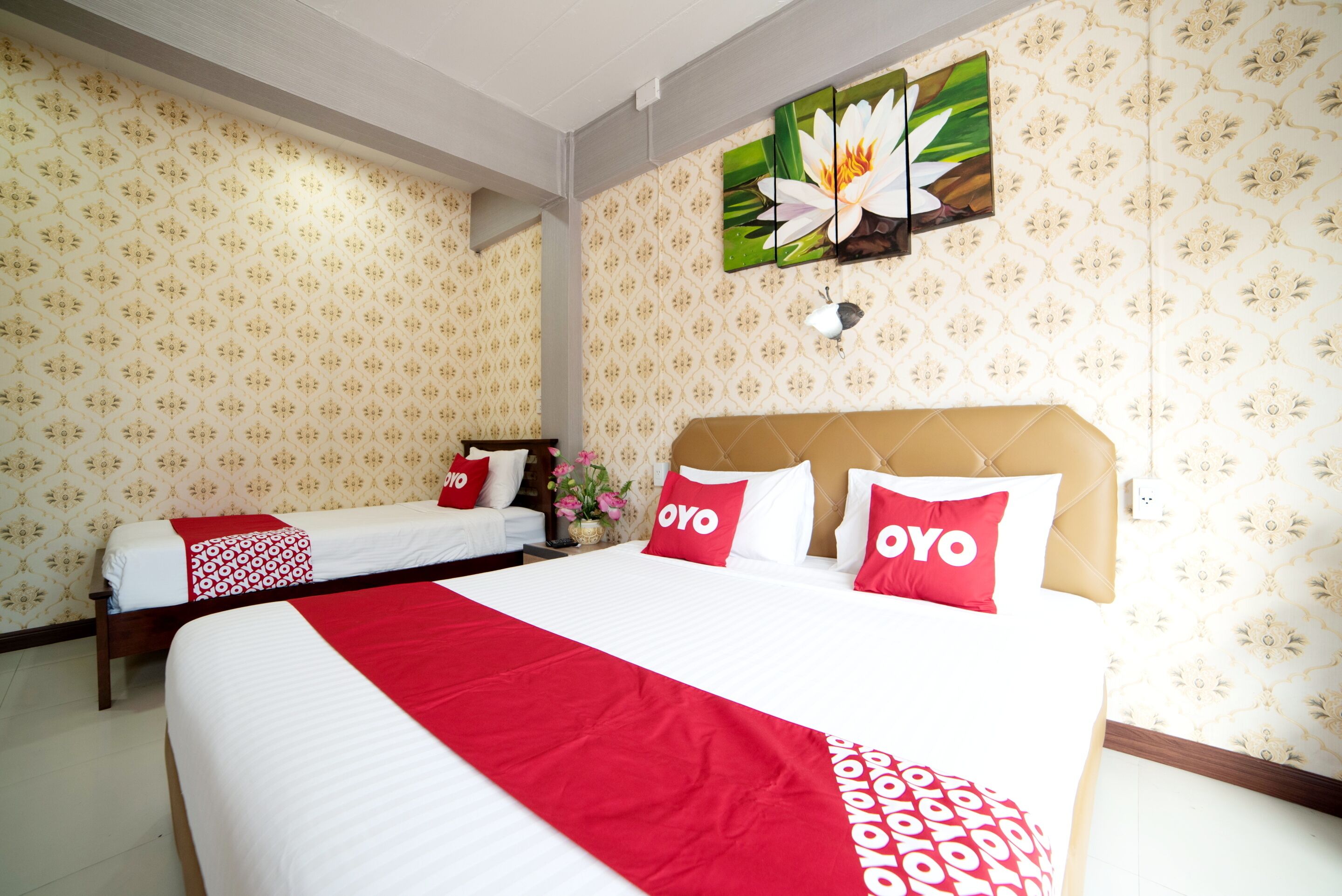 A hotel room with two beds, a large bed with red and white bedding, and a smaller bed with white bedding. There is a wall-mounted artwork of a flower, a vase with flowers, and a power outlet on the wall.