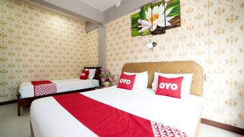 A hotel room with two beds, a large bed with red and white bedding, and a smaller bed with white bedding. There is a wall-mounted artwork of a flower, a vase with flowers, and a power outlet on the wall.